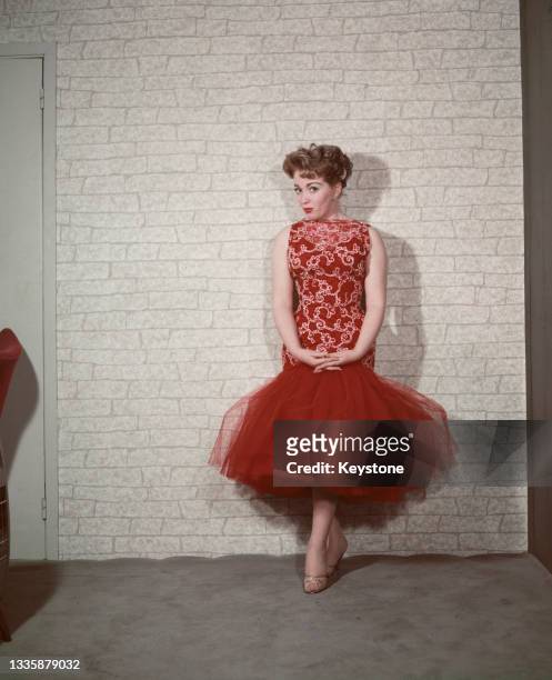 British singer Marion Ryan wearing a red-and-white dress with a red fishtail skirt, standing against a white brick wall backdrop, location...