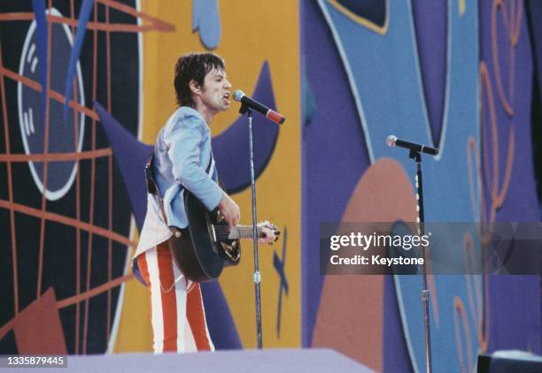 British singer, songwriter and musician Mick Jagger, wearing a blue jacket with red-and-white striped trousers, playing an Ovation acoustic guitar as...