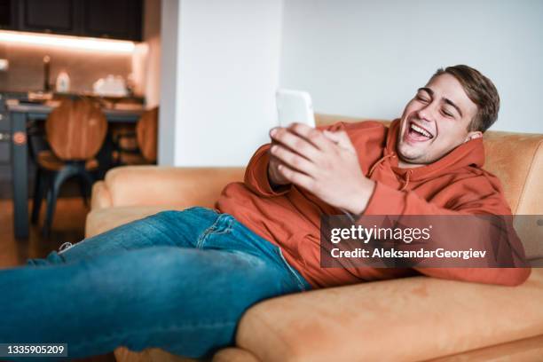 smiling male using smartphone and laughing while relaxing on sofa - lazy day stock pictures, royalty-free photos & images