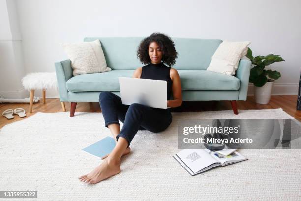 an african american millennial woman with curly hair, sits on the floor in the living room, as she works on her laptop, wearing a black top and black jeans. - african american woman barefoot stock pictures, royalty-free photos & images