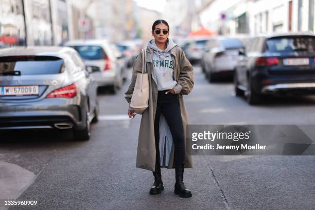 Sonia Dhillon wearing grey hoodie, olive trenchcoat, black denim and black Copenhagen boots and beige bag on August 03, 2021 in Munich, Germany.