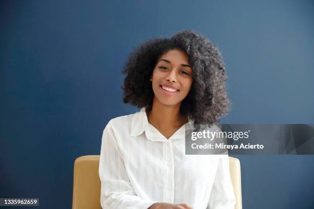 portrait of an african american millennial woman with curly hair, smiling in front of a dark blue background, while sitting in a camel colored leather chair, wearing a white button down top. - chairs in studio stockfoto's en -beelden