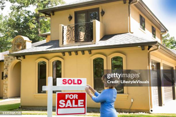 lovely young adult real estate agent standing beside her for sale sign in front yard of home.  she adds the sold sign and wears a blue top and jeans. - 拍賣 個照片及圖片檔