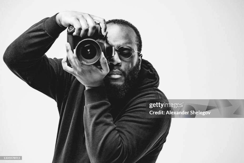 Professioneller Fotograf im Studio, Afroamerikaner tragen schwarzen Kapuzenpullover und Sonnenbrille mit Digitalkamera funktioniert. Schwarz-Weiß-Konzeptfotografie