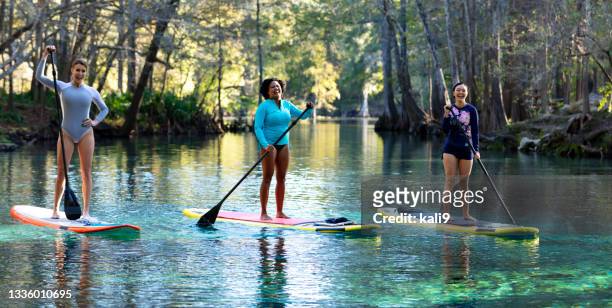 three women side by side paddle boarding on river, laugh - sup stockfoto's en -beelden