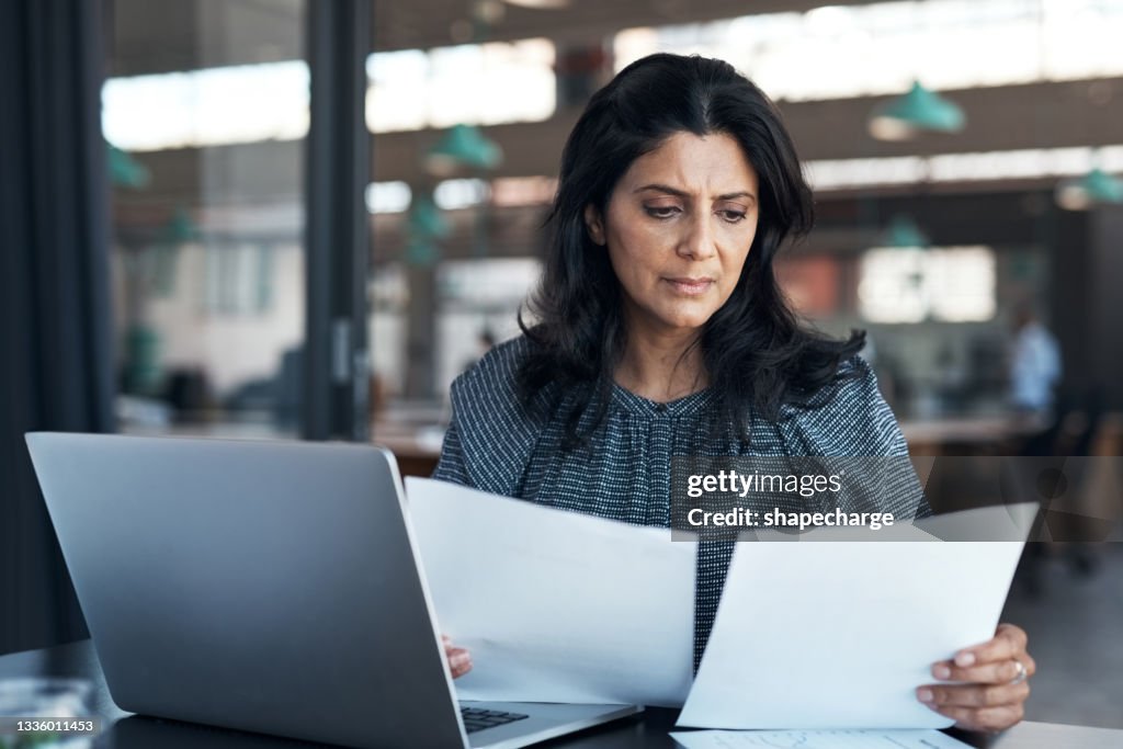 Shot of a mature businesswoman using a laptop and going through paperwork in a modern office