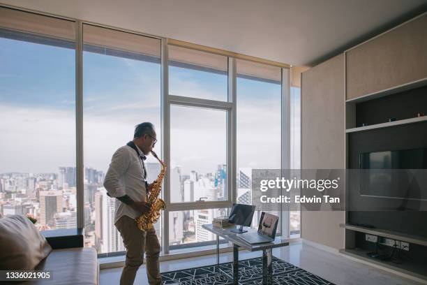 ライフハックアジアのアクティブなシニアマンアーティストは、サックスを演奏し、市内のアパートのリビングルームでラップトップを使用して彼の学生にそれを示しています - jazz showcase ストックフォトと画像