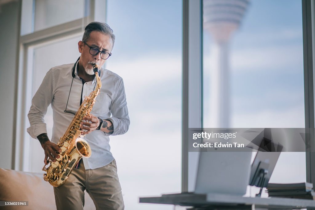 アジアのチネスアクティブシニア男性アーティストは、サックスを演奏し、市内のライブストリーミングでアパートのリビングルームでラップトップを使用して彼の学生にそれを示していま�