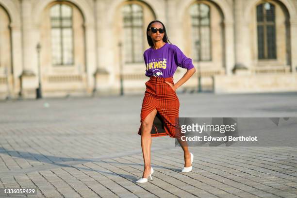 Emilie Joseph @in_fashionwetrust wears black large sunglasses, silver earrings, a purple with yellow and white print pattern logo and slogan...