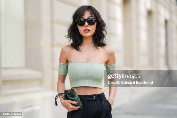 Xiayan wears black large sunglasses, silver earrings, a pale green ribbed off-shoulders cropped t-shirt, black high waist flowing wide leg pants, a...