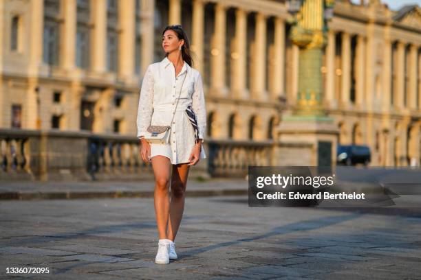 Amanda Derhy wears black sunglasses, pearls pendant earrings, a gold chain pendant necklace, a white cut-out and lace flower pattern buttoned / shirt...