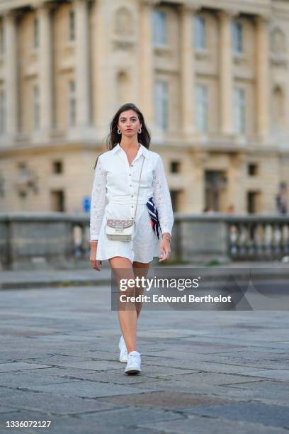 Amanda Derhy wears black sunglasses, pearls pendant earrings, a gold chain pendant necklace, a white cut-out and lace flower pattern buttoned / shirt...