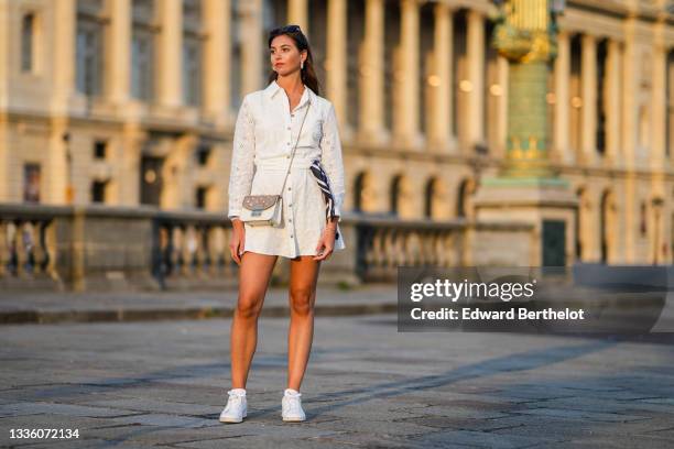 Amanda Derhy wears black sunglasses, pearls pendant earrings, a gold chain pendant necklace, a white cut-out and lace flower pattern buttoned / shirt...