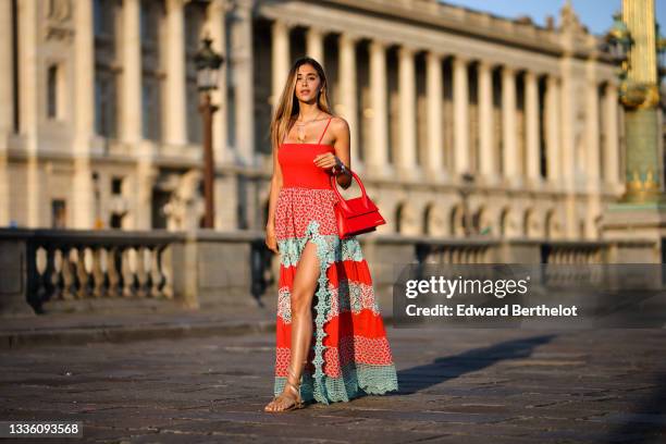 Jori Delli wears gold earrings, a gold chain necklace, silver chain pendants necklaces, a red ribbed V-neck / tank top with a long flowing slit /...