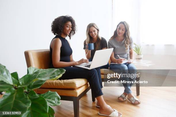 a multi-ethnic group of millennial women, sit together on leather chairs while working together using a laptop, in a brightly lit room wearing casual clothing. - brightly lit imagens e fotografias de stock