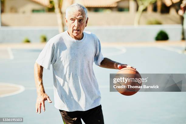 medium wide shot of senior man dribbling basketball during game on summer morning - septuagénaire photos et images de collection