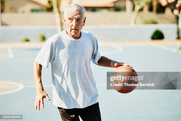 medium wide shot of senior man dribbling basketball during game on summer morning - anziani attivi foto e immagini stock