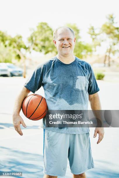 medium wide shot portrait of smiling mature male basketball player holding ball and standing on outdoor court on summer morning - pantalón corto gris fotografías e imágenes de stock