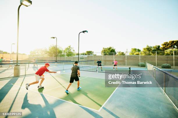 extreme wide shot of senior man hitting shot while playing doubles pickleball with friends on summer evening - 4 people playing games stock pictures, royalty-free photos & images