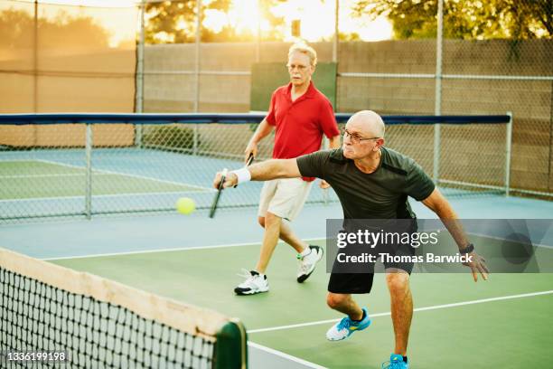 wide shot of senior man hitting backhand shot at net while playing doubles pickleball on summer evening - young at heart stock pictures, royalty-free photos & images