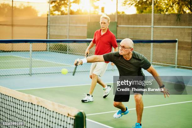 wide shot of senior man hitting backhand shot at net while playing doubles pickleball on summer evening - young at heart bildbanksfoton och bilder
