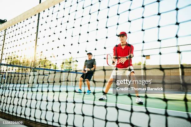 wide shot view through net of senior men playing doubles pickleball on summer evening - schlägersport stock-fotos und bilder