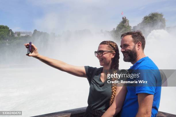 young couple taking selfie in front of rhine falls - chutes du rhin photos et images de collection