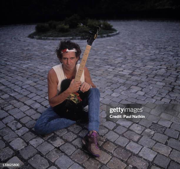 Portrait of British musician Keith Richards as he holds a guitar and sits on a sett-paved driveway, around the time of his 'Talk is Cheap' tour, late...