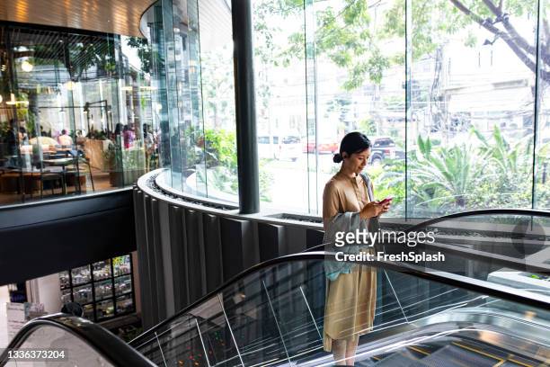 asian woman with a smartphone riding on an escalator - escalator stockfoto's en -beelden