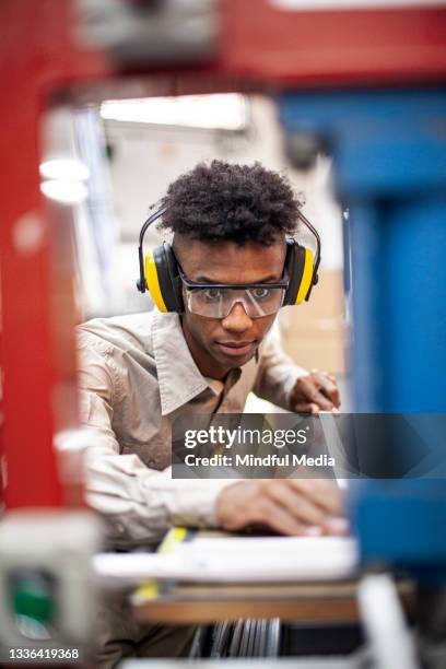 front view of male factory worker wearing protective earmuffs and plastic eyeglasses  next to industrial machinery - earmuff stock pictures, royalty-free photos & images