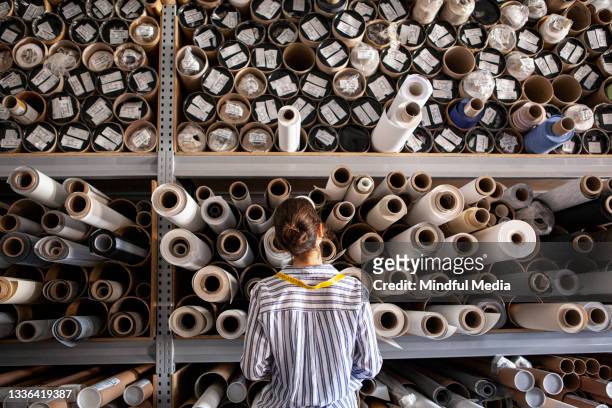 portrait of textile designer choosing fabric from stack of rolls inside sustainable workshop - fashion collection stockfoto's en -beelden