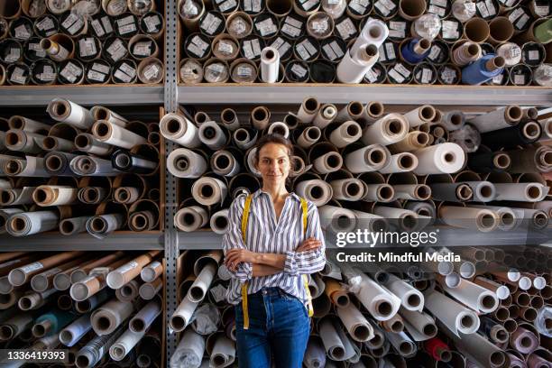 smiling young textile designer standing next to fabric rolls stacked in warehouse during daytime - textielfabriek stockfoto's en -beelden