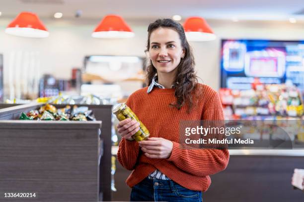 portrait of smiling young woman holding olives jar while standing inside filling station store - benzinestation stockfoto's en -beelden