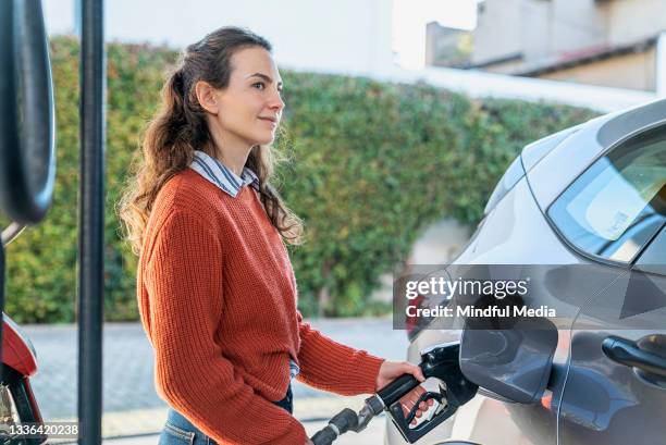 side view of young adult woman filling fuel tank while standing next to car - filling petrol stock pictures, royalty-free photos & images