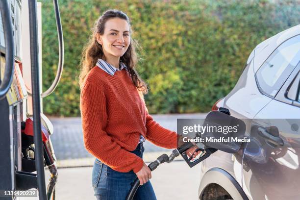 portrait of young adult woman filling fuel tank while standing next to car - filling petrol stock pictures, royalty-free photos & images