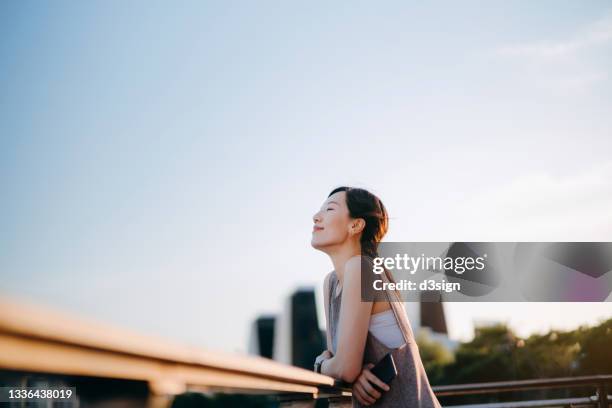 beautiful young asian woman with her eyes closed relaxing outdoors, setting herself free and feeling relieved. enjoying fresh air and the calmness with head up against sunlight at sunset - mulher chinesa imagens e fotografias de stock