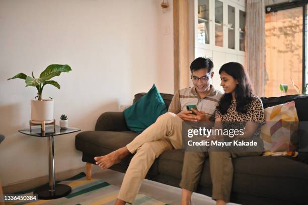 husband and wife sitting together on a sofa in living room and using phone - indian living room stock-fotos und bilder