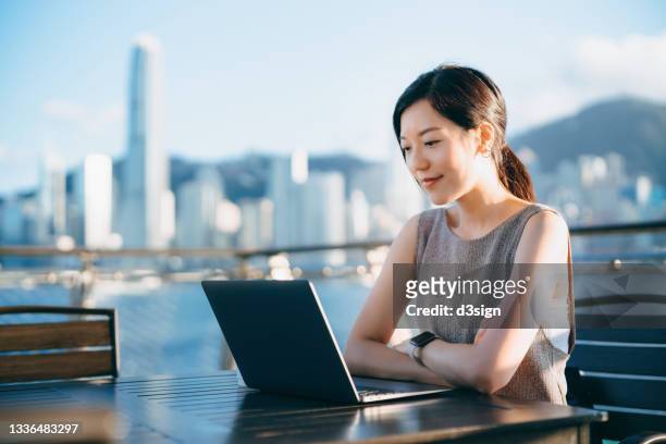 confident young asian businesswoman sitting outdoors by the promenade of victoria harbour, video conferencing on laptop with business partners, against urban cityscape in background. female leadership. remote working. business on the go - central asia stock-fotos und bilder