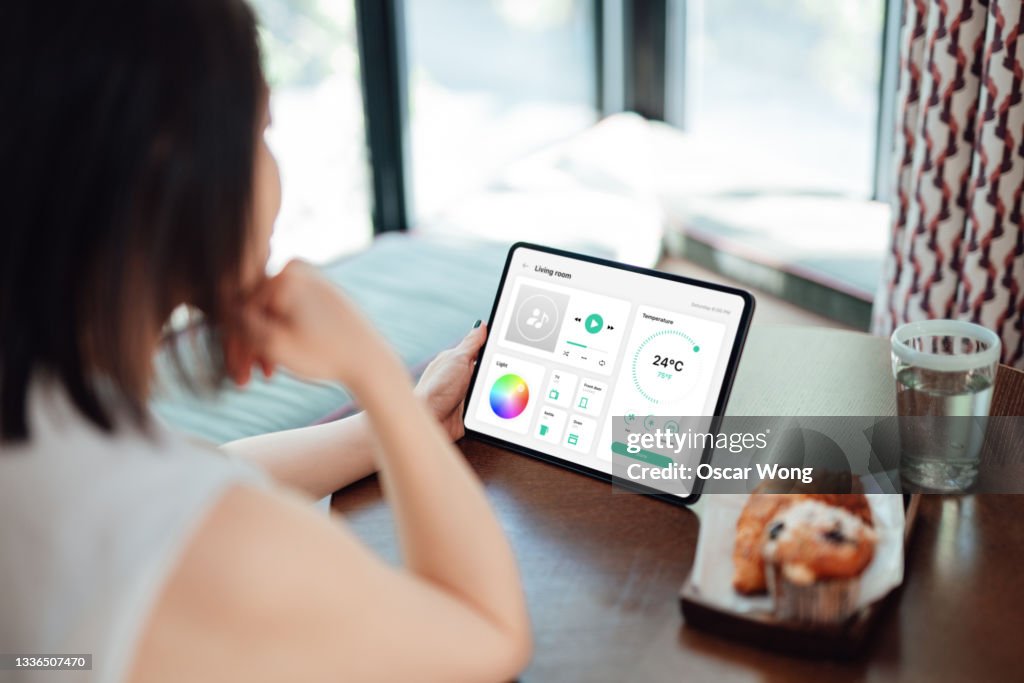Young woman using tablet to adjust the lighting equipment, control music, room temperature, oven, door lock, TV and kettle of a modern home