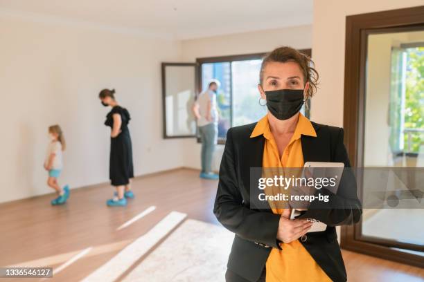 portrait of female real estate agent looking at camera and smiling with protective face mask while her clients looking around in new unfurnished house - businesswoman mask stock pictures, royalty-free photos & images