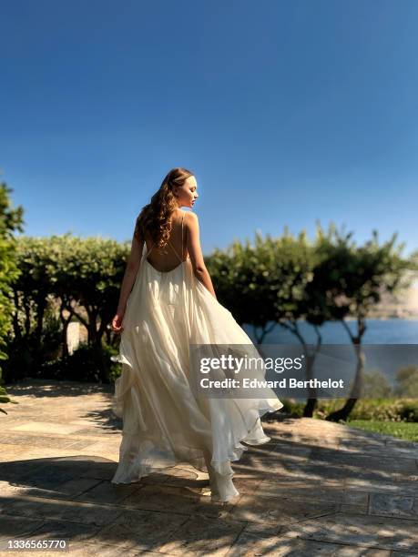 Model Cristina Padure wears a white long pleated/gathered evening/wedding sleeveless back-nude and off-shoulder dress from Revelle, high heels...