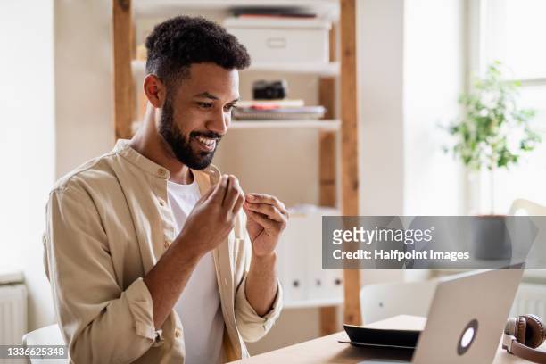 young man with laptop using sign language indoors in office, video call concept. - sign stock-fotos und bilder