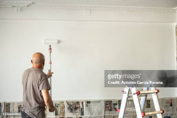 a senior painter seen from behind while applying white paint with a roller - white paint roller stock pictures, royalty-free photos & images