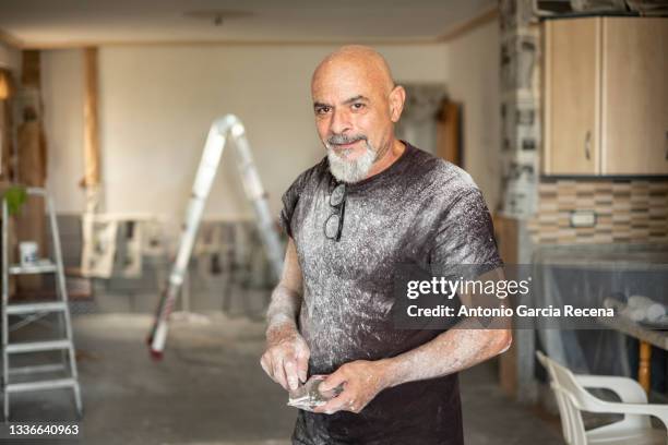 a senior bricklayer poses in portrait with tools in hand smiling and looking at camera, full of paint and plaster scrapes - arab old man fotografías e imágenes de stock