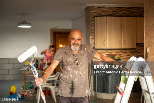 a senior wall painter poses in portrait with tools in hand smiling and looking at camera, full of paint with a roller - casa real española fotografías e imágenes de stock