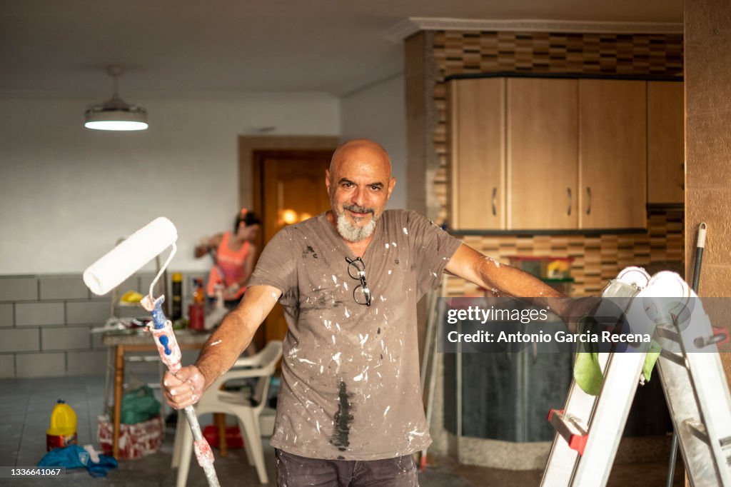 A senior wall painter poses in portrait with tools in hand smiling and looking at camera, full of paint with a roller