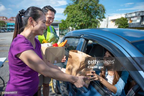 young volunteers cheerfully hands a paper bag full of groceries for car driver through the car window. - food distribution stock pictures, royalty-free photos & images