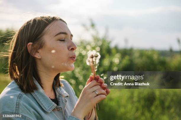 young woman blowing dandelion seed in meadow outdoors in sunny day. vibrant summer portrait. - spring foto e immagini stock