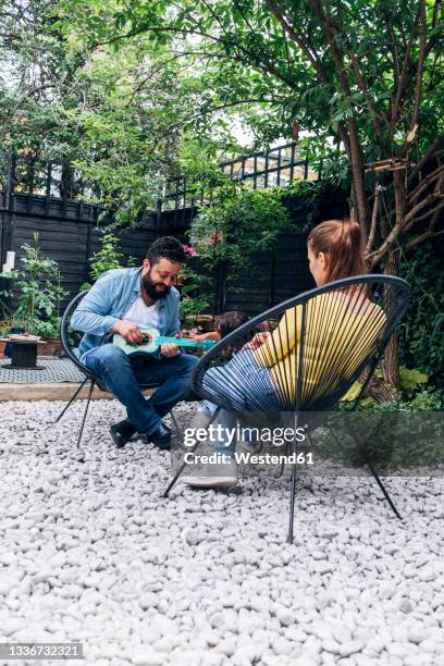 father playing guitar while sitting with family in back yard - family back yard stockfoto's en -beelden