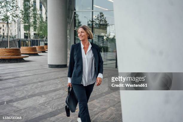 smiling female professional with bag looking away while walking on walkway - tre quarti foto e immagini stock